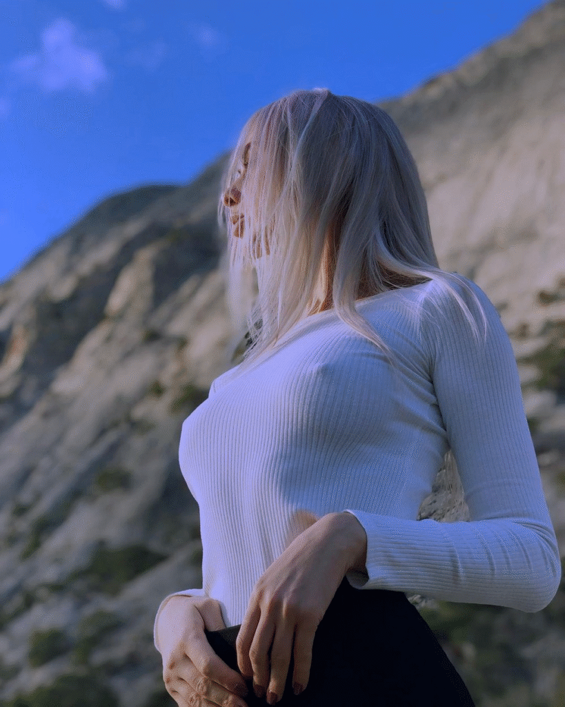Mulher loira, jovem modelo, com cabelo esvoaçante com blusa branca justa, ao ar livre em paisagem montanhosa e céu azul