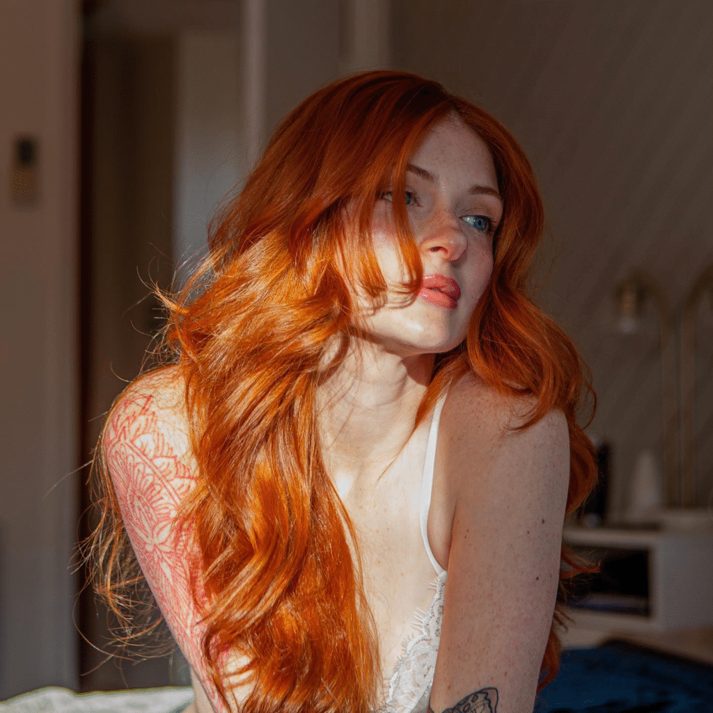 Sexy Redheads: A Beleza do Rutilismo. Mulher ruiva com longos cabelos ondulados e olhos claros, sentada com a luz do sol realçando suas sardas. Veste uma lingerie de renda branca e exibe tatuagens nos braços. A imagem tem um tom sensual e artístico, capturando a beleza feminina com luz natural.
