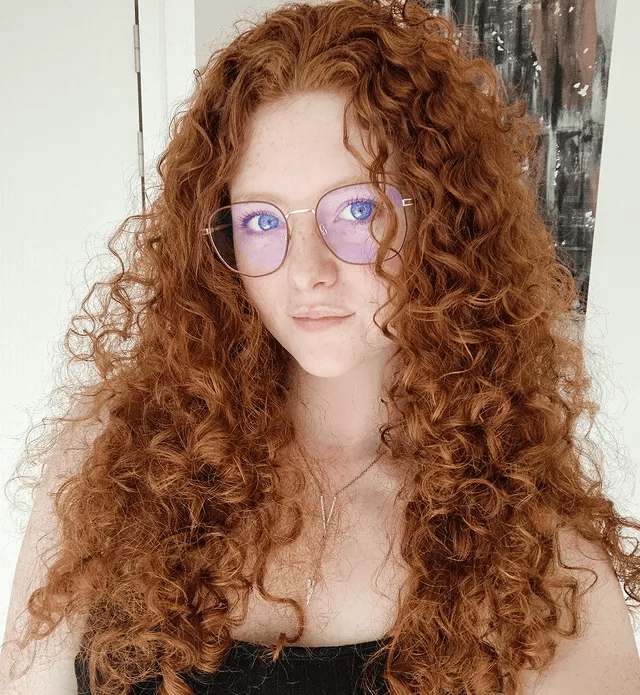 Sexy Redheads: A Beleza do Rutilismo. Retrato em close-up de uma mulher ruiva com cabelos cacheados e volumosos. Ela tem olhos azuis brilhantes, sardas e usa óculos modernos. A imagem destaca a beleza natural e o contraste entre o cabelo ruivo e os olhos claros.