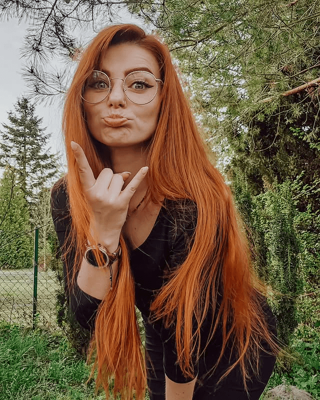 Sexy Redheads: A Beleza do Rutilismo. Mulher ruiva com cabelos longos e lisos, usando óculos redondos. Ela faz um gesto de "rock and roll" com a mão e uma expressão divertida com a boca. A imagem é em ambiente externo, com vegetação e uma cerca ao fundo.