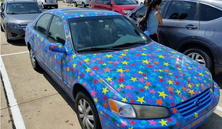 Carro de cor prata estacionado com a lataria totalmente coberta por adesivo azul vibrante cheio de estrelas coloridas nas cores vermelho, amarelo, verde e roxo, destacando-se entre outros veículos de cores convencionais em um estacionamento.