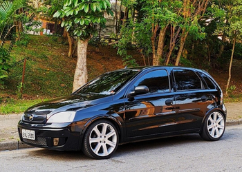 carro Chevrolet Corsa hatchback de segunda geração, na cor preta e com pintura brilhante. O veículo está estacionado de perfil em uma rua de asfalto, possui vidros totalmente escurecidos (insulfilm) e rodas de liga leve esportivas prateadas de tamanho grande.