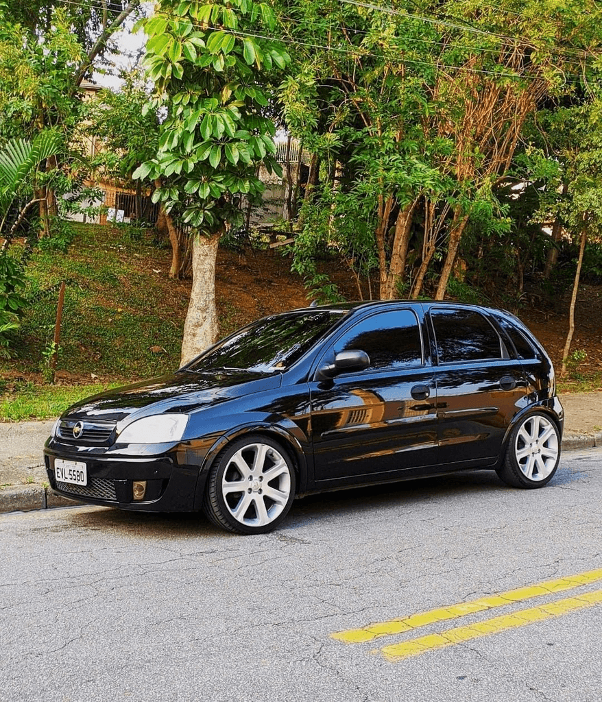 carro Chevrolet Corsa hatchback de segunda geração, na cor preta e com pintura brilhante. O veículo está estacionado de perfil em uma rua de asfalto, possui vidros totalmente escurecidos (insulfilm) e rodas de liga leve esportivas prateadas de tamanho grande.