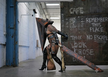 Full-shot of a person cosplaying a gender-bent Female Pyramid Head from Silent Hill in a gritty, industrial corridor. They are wearing high-heeled boots, leather straps, and carrying the iconic massive Great Knife while posing next to a rusted metal door with graffiti.